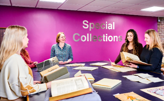 3 girls smiling gathered around a large table with Special Collections documents on top, taking part in a Special Collections teaching session, one girl looking through a book. In front of a purple wall with 'Special Collections' in silver on the wall